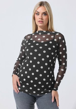 Kvinde iført en sort gennemsigtig langærmet top med store hvide polkaprikker over en sort camisole, kombineret med grå jeans.