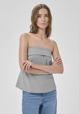 Žena so svetlohnedými vlasmi nosí ramienkový sivý top a modré rifle, stojí so skríženými rukami pred jednoduchým pozadím.