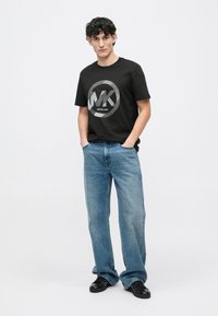 T-shirt preta de mangas curtas com logótipo branco e design circular, combinada com jeans de corte largo em azul claro e sapatos pretos.