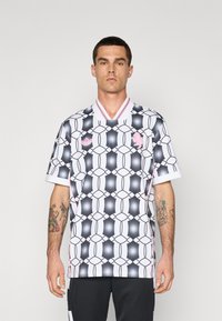 Jersey blanco de manga corta con un patrón geométrico en negro y rosa, con escote en V y ribete de color contrastante en el cuello. Logo de la marca deportiva en el pecho.