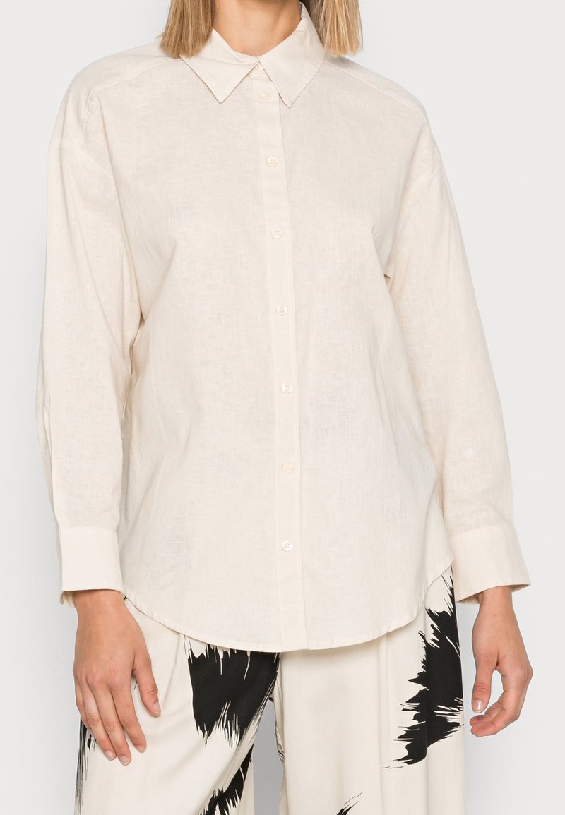 Femme portant une chemise crème à manches longues avec boutons, associée à un pantalon blanc orné de motifs abstraits en éclaboussures de peinture noire.