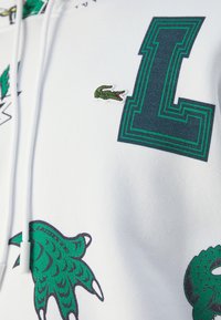 Sweat à capuche blanc avec grande lettre "L" en vert et noir, petit logo crocodile vert et motifs graphiques verts de griffes et de queue.