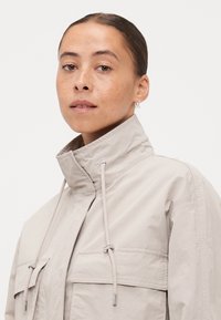 Light beige jacket with a high collar, drawstring closure, and two front pockets. Made of smooth, lightweight fabric with a relaxed fit.