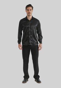 Chemise noire en satin à manches longues avec un devant à boutons, col classique et texture lisse, associée à un pantalon noir et des chaussures.