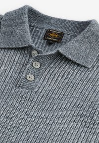 Camisa polo de punto gris con textura acanalada, tres botones y una etiqueta con el texto "Bienes Duraderos de Grado Premium 1982."