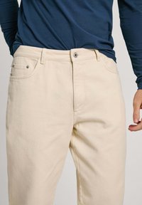Pantalones de algodón beige con un diseño de pierna recta, que cuentan con bolsillos delanteros, cierre de botón y detalles de costura visibles.