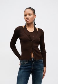 Rjava rebrasta jopica z globokim izrezom, zapenjanjem na gumbe in dolgimi rokavi. Nosena preko ujemajoče se majice, kombinirana s temno modrimi jeans hlačami.