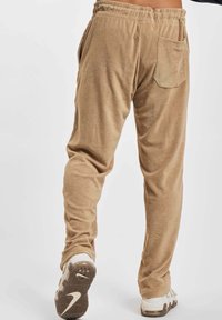 Person wearing tan terry cloth pants with elastic waistband and back pocket, shown from behind, paired with white sneakers.