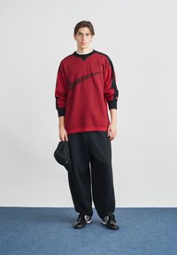 Piros-fekete csíkos kötött pulóver hosszú ujjakkal, laza fekete nadrággal és fekete tornacipővel párosítva. Bal kézben tartott fekete sapka.