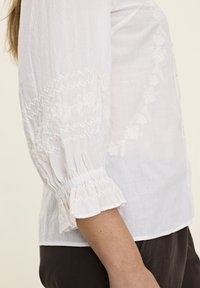 Blouse blanche avec des manches longues légèrement bouffantes, des poignets élastiques et de délicates broderies florales le long des manches et du corps.