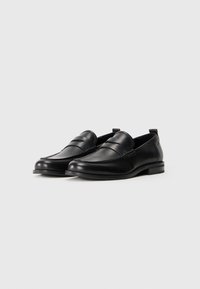Et par svarte skinn penny loafers med avrundede tær og lave hæler på en hvit bakgrunn.