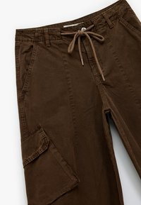 Brązowe bawełniane spodnie cargo z elastycznym pasem na sznurek, bocznymi kieszeniami i pojedynczą kieszenią cargo, charakteryzujące się miękką fakturą i luźnym krojem.