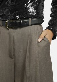 Persoon in taupe broek met hand in de zak, zwarte leren riem met gouden gesp, en zwart pailletten top.