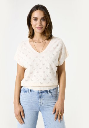 Strikket cremefarvet v-hals top med korte ærmer, et tekstureret bladmønster og en ribbestrikket kant. Sammen med lyseblå jeans.