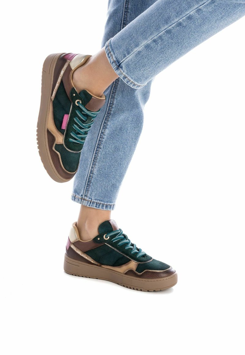 Zapatillas de ante verdes y marrones con cordones turquesas, acentos beige y una suela gruesa color tostado. Presenta un detalle rosa en el talón.