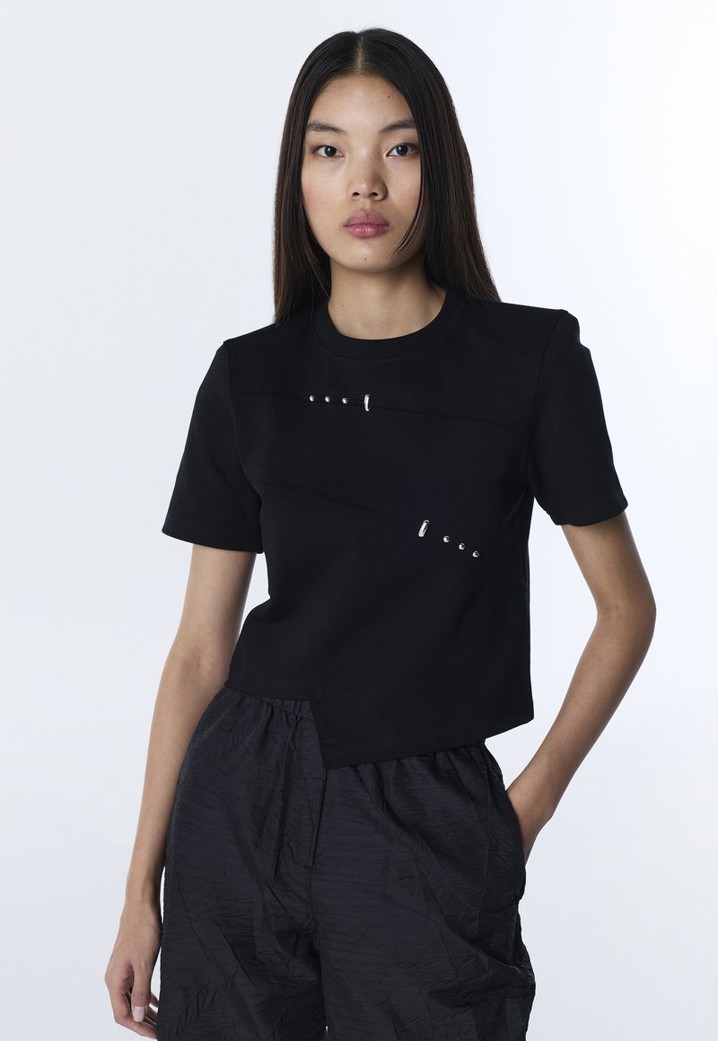 Jeune femme aux cheveux longs et raides portant un haut noir à manches courtes avec des clous argentés et un pantalon noir texturé, debout avec une main dans la poche.