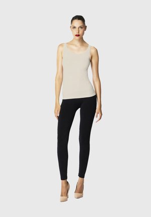 Donna con capelli scuri con canottiera beige, leggings neri e tacchi alti beige, in piedi davanti a uno sfondo chiaro e uniforme.