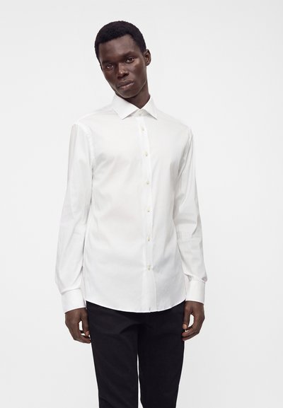 Uomo che indossa una camicia bianca a maniche lunghe abbottonata e pantaloni neri, in piedi davanti a uno sfondo chiaro e semplice.