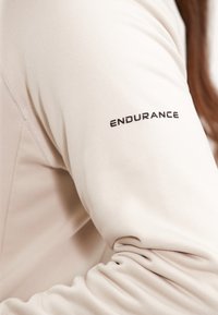 Ljusbeige långärmad sporttröja med slät textur; har svart "ENDURANCE"-text på vänster ärm. Figurpassad design med subtila sömdetaljer.
