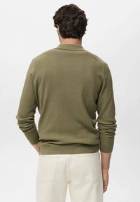 Pull en tricot côtelé vert olive avec des manches longues et un col rond, associé à un pantalon clair avec des poches arrière.