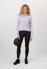 Moteris su garbanotais plaukais, apsirengusi šviesiai violetinėje užtraukiamoje striukėje ir juodose dviratininkiškose tamprėse, laiko juodą dviratininkų šalmą, stovėdama ant vientiso balto fono.