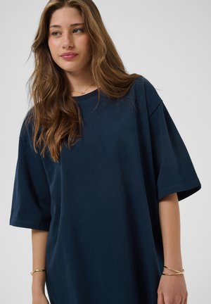Jeune femme aux longs cheveux châtain clair portant un t-shirt bleu marine surdimensionné et des bracelets dorés sur fond uni.