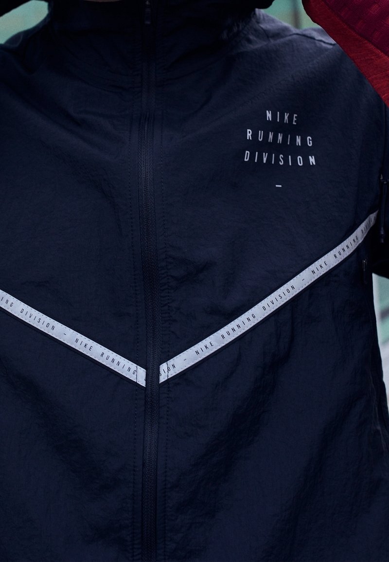 Gros plan d'une veste de course noire Nike avec une bande réfléchissante diagonale et le texte "Nike Running Division" sur la poitrine et la bande.