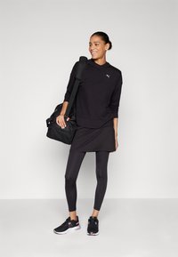 Sweatshirt de sport noir avec capuche, superposé à une jupe noire, associé à des leggings noirs et des baskets. Sac de sport noir en bandoulière.