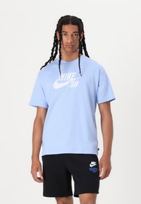 Gaiši zils Nike SB T-krekls ar baltu logo priekšpusē, apvienots ar melniem īsiem šortiem ar zilu Nike zīmolu. Kokvilnas materiāls, brīvā piegriezuma.