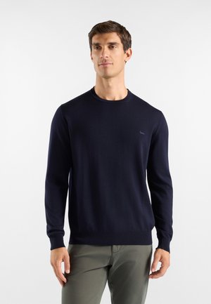 Uomo che indossa un maglione girocollo blu scuro semplice e pantaloni verde oliva, in piedi contro uno sfondo bianco.