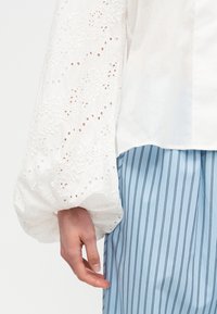 Χέρι με λευκό δαντελένιο μανίκι μπαλόνι και μπλε φούστα με ρίγες σε απλό φόντο.