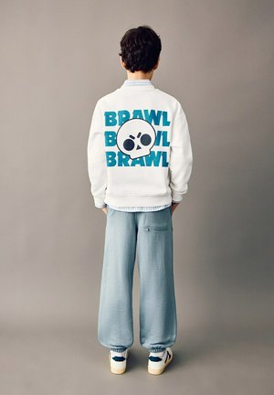 Băiat stă cu fața spre spate purtând o geacă albă cu text albastru „BRAWL” și o grafică mare cu craniu, pantaloni albastru deschis și adidași albi.