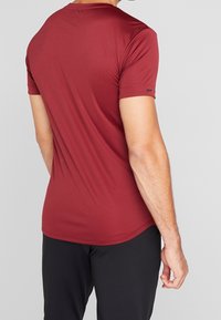 Maroon kortärmad träningsskjorta i slät, lättviktsmaterial. Har en rundad halsringning och en diskret logotyp på ärmen.