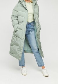 Pitkä, pehmeä takki vaaleanvihreänä, jossa on huppu, vetskari, sivutaskut ja tikattu pinta. Yhdistetty sinisiin farkkuihin ja valkoisiin lenkkareihin.