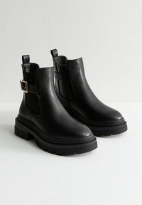 Botas de cuero negro hasta el tobillo con suelas gruesas, cremallera lateral en una bota y correa con hebilla en la otra, colocadas sobre una superficie blanca.