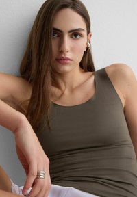 Mujer joven con cabello largo y castaño, vestida con un top verde olivo y pendientes plateados, sentada frente a una pared gris claro, mirando directamente hacia adelante.