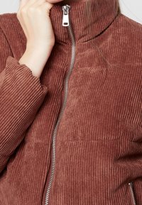 Hnědá kordurová bunda s vysokým límcem, se stříbrným zipem a žebrovanou texturou na rukávech. Detailní záběr, který ukazuje ruku poblíž límce.