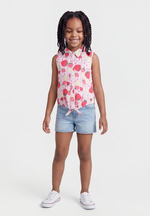 Mosolygó lány fonott hajjal, ujjatlan rózsaszín pólót visel, amelyen eperminták vannak, világos farmer rövidnadrágot és fehér sportcipőt.