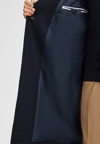 Hand die een open donkerblauwe jas vasthoudt met een gladde satijnen binnenvoering en een gestikt zakje, gedragen over camel-kleurige broeken.