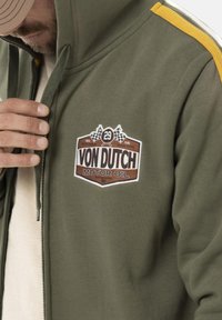Sweat à zip olive avec des rayures jaunes et blanches sur les épaules, arborant un logo "Von Dutch Motor Oil" marron et blanc sur la poitrine.