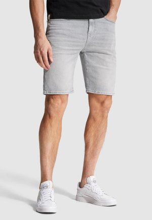 Homme portant un short en jean gris clair, une chemise noire et des baskets blanches, une main dans la poche, debout devant un fond uni.