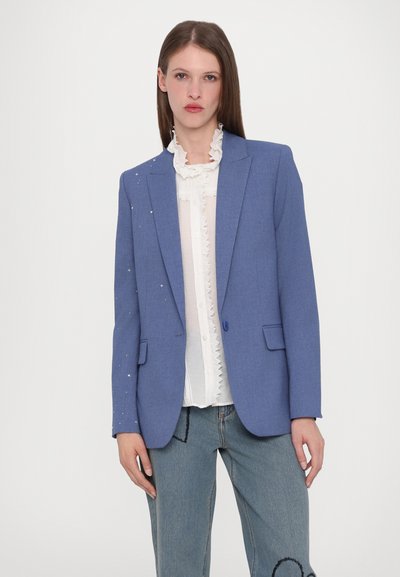 Blauwe blazer van textuurstof, met één knoopsluiting, twee voorzakken, versierd met kleine kraaltjes op één mouw, gestyled over een witte blouse.