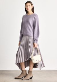 Moteris dėvi levandų spalvos megztinį ir klostuotą alyvinės spalvos sijoną, laiko smėlio spalvos rankinę ir dėvi taupe spalvos aulinukus, stovėdama paprastoje fono aplinkoje.