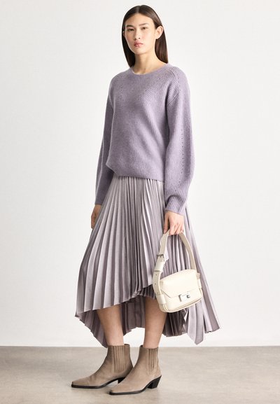 Femme portant un pull lavande et une jupe plissée lilas, tenant un sac à main beige et chaussée de bottines taupe, debout sur un fond uni.