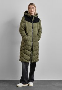 Lång olivgrön puffjacka med svart axelparti, vertikalt kviltad design, dragkedja fram, sidofickor och en justerbar huva.