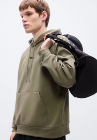 Φούτερ με κουκούλα σε ελιά πράσινο χρώμα με μπροστινή τσέπη και κορδόνι στην κουκούλα, σε συνδυασμό με μια μαύρη τσάντα σε στρογγυλό σχήμα και με στιβαρό λουράκι.