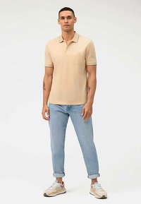 Beige poloshirt met een kraag en korte mouwen, met een borstzakje. Gecombineerd met lichtblauwe spijkerbroek en beige sneakers.