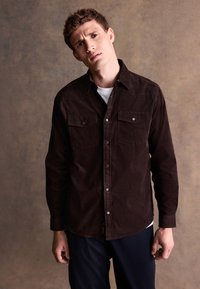 Brązowa koszula sztruksowa z kołnierzykiem na guziki, dwoma kieszeniami na piersi i zatrzaskami, noszona na białej koszuli, na neutralnym tle.