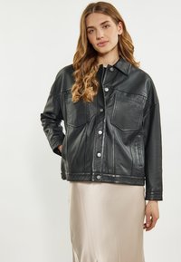 Jachetă din piele neagră cu închidere prin nasturi, două buzunare pe piept și o croială lejeră. Textură netedă și design clasic al gulerului.