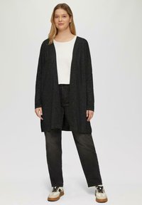 Schwarze Strickjacke mit lockerer Passform, offener Vorderseite und dezenter Textur. Kombiniert mit dunklen Jeans und beigen Sneakers mit schwarzen Akzenten.